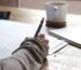 person writing on brown wooden table near white ceramic mug